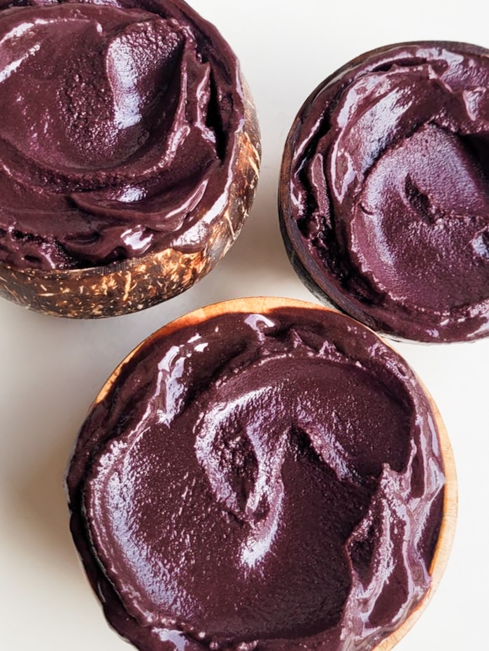 Trois bols remplis d'açaí épais et violet foncé, deux dans des coques de noix de coco et un dans un bol en bois clair, sur fond blanc.