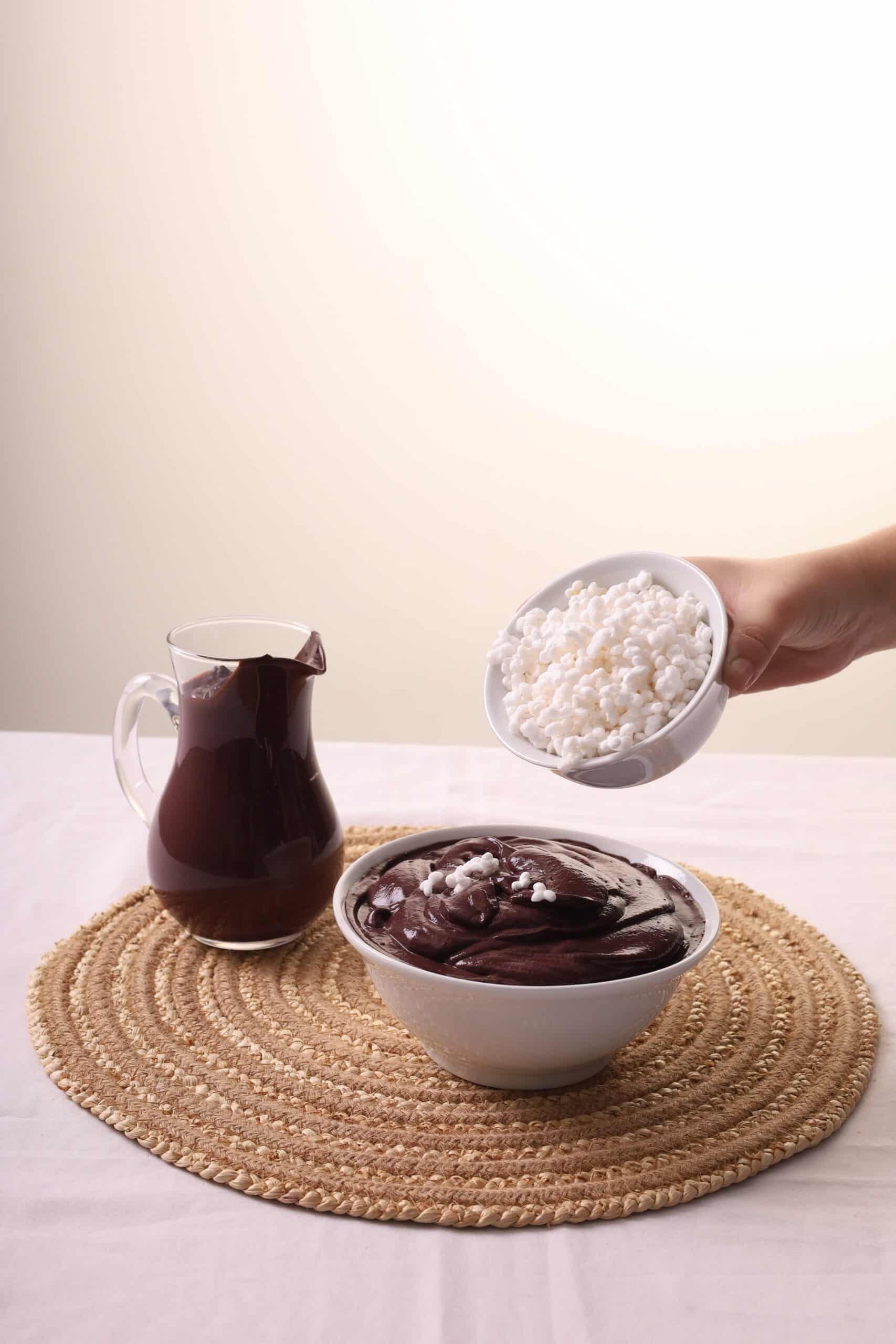 Main ajoutant des granulés blancs (tapioca/sago) sur un bol d'açaí épais et sombre, à côté d'une carafe du même mélange.