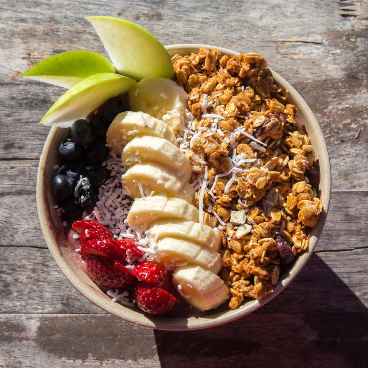 Bol d'açaï ou smoothie bowl garni de granola croustillante, tranches de banane, fraises, myrtilles, copeaux de noix de coco et quartiers de pomme verte.