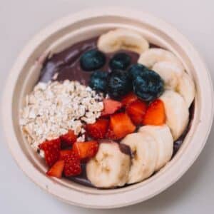 Bol d'açaï épais et violet garni de bleuets, fraises coupées, rondelles de banane et flocons d'avoine sur fond clair.