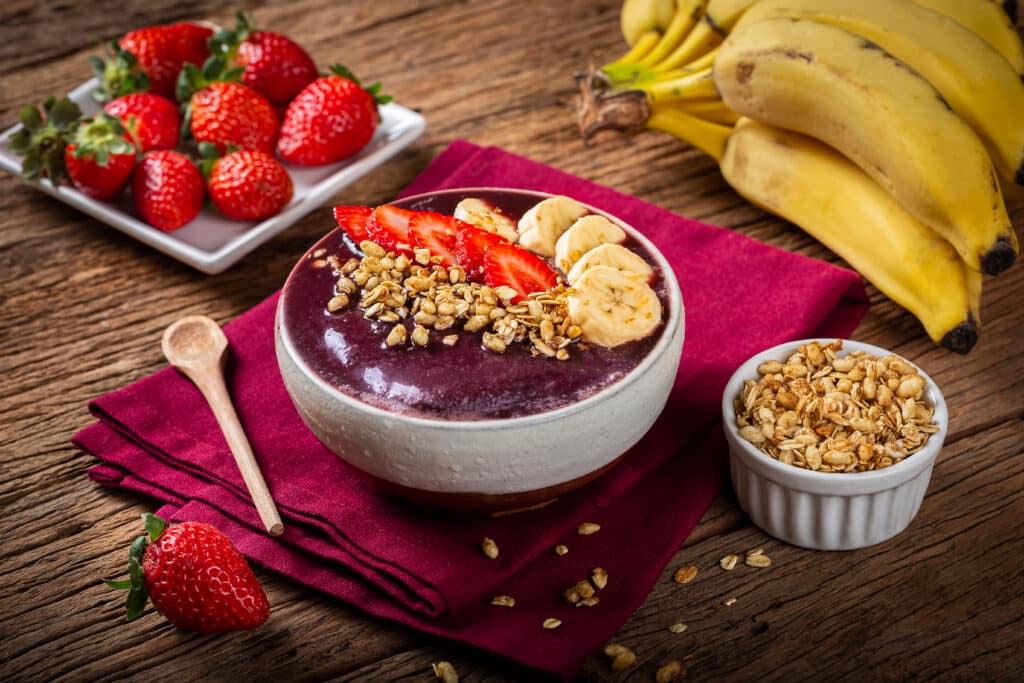 Açaí Bowl Frais avec Fruits et Granola sur Bois Bol d'açaí violet épais garni de tranches de fraises, rondelles de banane et granola, sur une serviette bordeaux, avec bananes et fraises à côté.