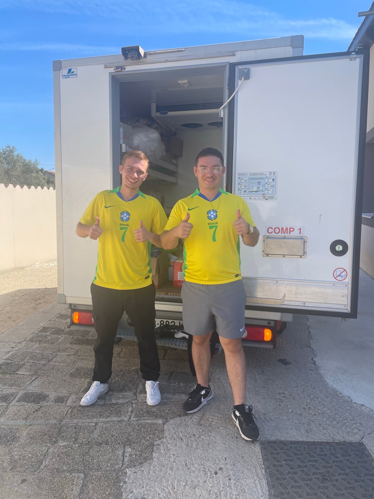 Deux hommes souriants portant des maillots jaunes du Brésil avec le numéro 7, pouces levés devant une camionnette frigorifique ouverte avec l'inscription COMP 1.